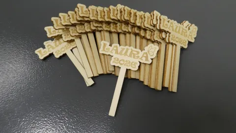 A fan-shaped stack of matching custom wooden cupcake toppers precisely engraved with Laura 2026 on a dark surface.